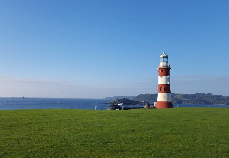 Scenic view of Smeaton's Tower lighthouse by the sea with clear blue skies and grassy foreground.